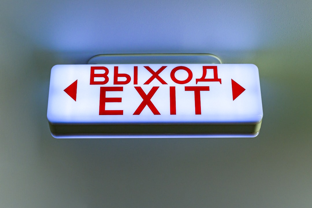 A glowing bilingual exit sign in Russian and English hangs from the ceiling, lit in a cool blue hue. The bold red text “ВЫХОД / EXIT” with directional arrows stands out sharply, reflecting a blend of utilitarian design and international signage standards.